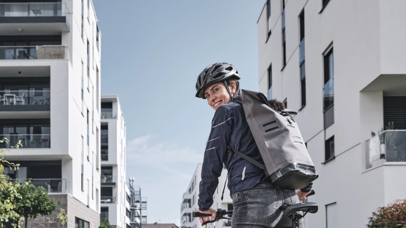Eine Fahrradfahrerin fährt und schaut lächeln über die Schulter zurück. Sie trägt eine blaue Jacke und einen grauen Ruckssack.